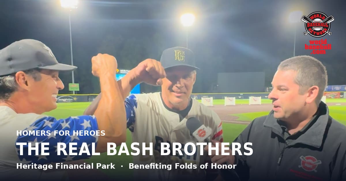Former major leaguers Jose and Ozzie Canseco, one in a cream Bash Brothers jersey and one in a gray cap, bump forearms on a lit ballfield at night beside a reporter holding a World Baseball Network microphone.