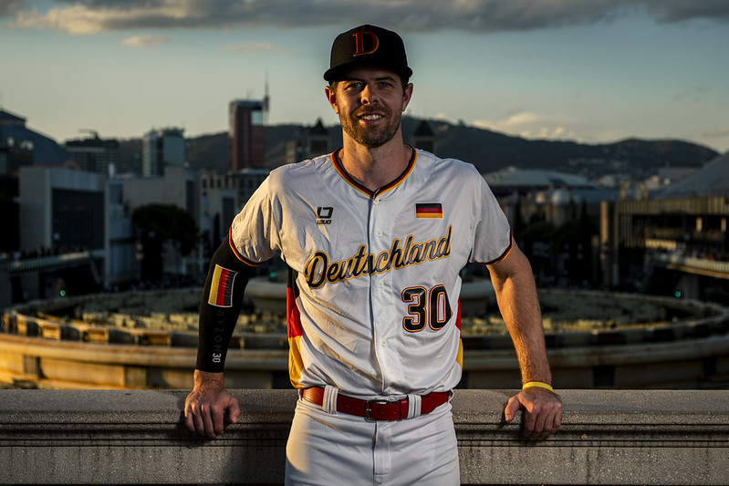Germany infielder and captain Eric Brenk in the team’s Deutschland uniform, posed on a terrace with the Barcelona skyline behind him at sunset.